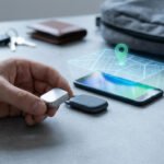 Main tenant une balise de suivi argentée près d'un smartphone affichant une carte holographique avec une épingle de localisation. Clés, portefeuille et sac à dos en arrière-plan.